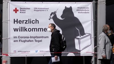 Två män i munskydd går på gatan i Berlin. I bakgrunden syns en reklamskylt för coronavaccin.