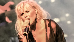 Josefin Nilsson en kvinna med blont hår och svarta kläder står på en scen och sjunger i en mikrofon, i bakgrunden syns en gitarrist.