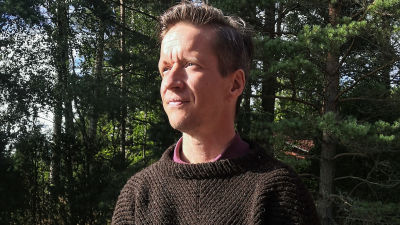 Markus har en brun stickad tröja på sig. Han sitter ute med granar i bakgrunden.