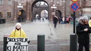 Greta Thunberg. På bilden syns klimataktivisten i ett regnigt Stockholm. Hon är klädd en gul regnjacka och håller i sin kända skylt "Skolstrejk för klimatet".