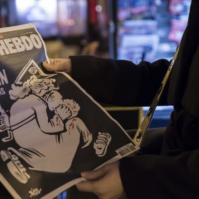 Charlie Hebdo-lehden erikoisnumerossa muistetaan vuoden takaista terrori-iskua. Lehden etukannessa parrakas jumala, jonka selässä rynnäkkökivääri.