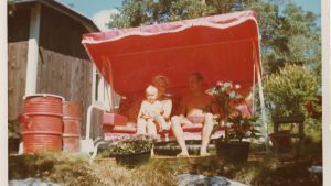 Helena Ahti-Hallberg alle 2-vuotiaana vanhempiensa kanssa aurinkoisella mökkipihalla puutarhakeinussa.