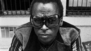 Miles Davis poseeraa kädet ristissä, ranteessa iso kultaketju ja silmillä suuret aurinkolasit