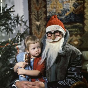 Pikkutyttö joulupukin sylissä jouluna. Takana joulukuusi ja seinällä ryijy.