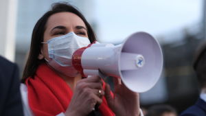 En av den belarusiska oppositionens främsta gestalter är Svetlana Tichanovskaja. Den senaste tiden har hon träffat europeiska flera höjdare. Här är hon klädd vitt och rött, precis samma färger som flaggan som demonstranterna nu har anammat som sin symbol. Svetlana Tichanovskaja talar i en megafon. Hon har munskydd på sig och en röd halsduk runt halsen.