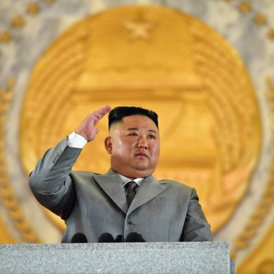 Kim Jong-un seisoo puhujanpöntön takana harmaassa puvussa ja tervehtii paraatia.