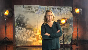 Elli Toivoniemi. Elli Toivoniemi seisoo studiossa taustalavasteen ja studiolamppujen edessä. Kuva otettu lyhytvideosarjaan Me elokuvantekijät.