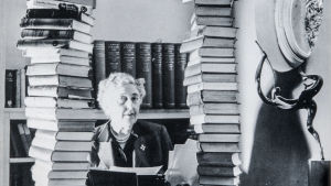 Agatha Christie - rikosten kuningatar. Agatha Christie katsoo kameraan kahden korkean kirjapinon välistä.