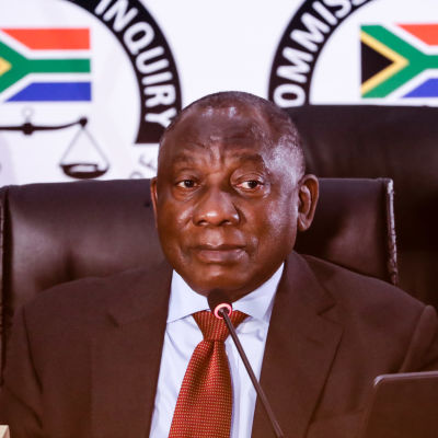 Sydafrikas president Cyril Ramaphosa sitter på en stol och tittar åt sidan. Han är klädd i kostym och har en mikron framför sig på ett bord. I bakgrunden syns Sydafrikas flagga på väggen.