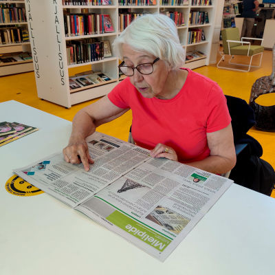 En dam i röd skjorta sitter vid ett bord i ett bibliotek och läser en tidning.