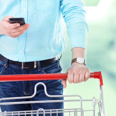 Mies työntää ostoskärryä älypuhelin kädessä