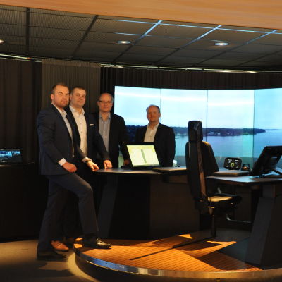 Fyra herrar står kring bordet vid Kongsbergs fartygssimulator, som föreställer ett bord från vilket man kan fjärrstyra ett fartyg.