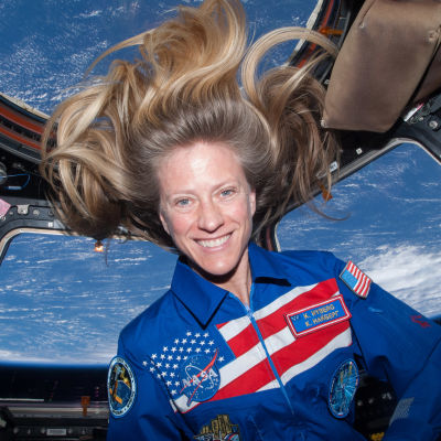 Astronautti Karen Nyberg katsoo kameraan hymyillen avaruussukkulassa, jonka ikkunoista näkyy maapallo.