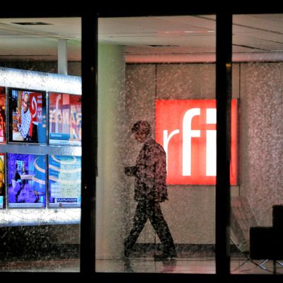 Kuva RFI:n aulasta. Mies kävelee RFI:n punavalkoisen logon ohi. Vasemmalla näkyy televisionäyttöjä, joissa on meneillään uutislähetyksiä. Oikeassa reunassa näkyy France24-uutiskanavan logo.