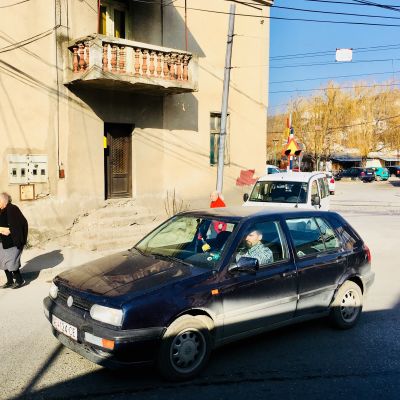 En liten och gammal, smutsig bil kör förbi en äldre dam i den makedonska staden Veles.