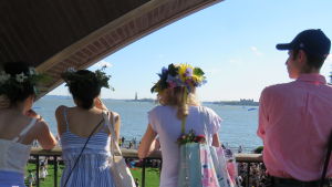 Frihetsstatyn och Ellis Island i bakgrunden. Kvinnor med blomsterkransar på huvudet tittar ut över midsommarfirande och frihetsstatyn i New York.