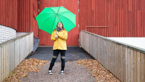 Anne Kassimäki seisoo Navetta-teatterin seinustalla vihreän sateenvarjon alla.