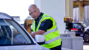 Ett sällskap på tre bilar har kommit från Sverige för att ha semester på Åland och blir både förvånade och upprörda när sjöbevakaren Mats berättar om de inreserestriktioner som finns. En manlig sjöbevakare står i gul väst viden en bil och pratar med någon inne i bilen.
