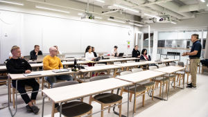 Vid Aalto-universitetet kunde studerande välja om de deltog i undervisningen på distans eller på plats. Universitetet avråder ändå från att studera utomlands den här hösten och ser gärna att internationella studerande i Finland studerar på distans. Ett tiotal studerande sitter glest i en föreläsningssal. Till vänster sitter en man i gul tröja, till höger står läraren.