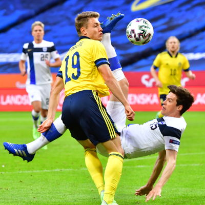 Finländsk fotbollsspelare i vitt ligger i luften och sparkar bort bollen framför svensk spelare i gult.
