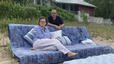 Ett par sitter i en soffa på en sandstrand med en sommarstuga i bakgrunden.