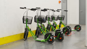 Sparkcyklar är ett bra hjälpmedel för dem i personalen som behöver ta sig från en arbetspunkt till en annan. Annars skulle det bli många långa kilometrar att gå per dag. flera gröna sparkcyklar på rad mot en vit vägg.