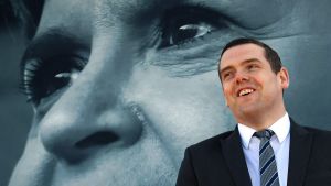 Den konservative Toryledaren Douglas Ross i Skottland kritiserar Sturgeon för att fokusera på skotsk självständighet medan pandemin pågår. Konservativa partiets ledare i Skottland Douglas Ross med en plansch på Nicola Sturgeon i bakgrunden