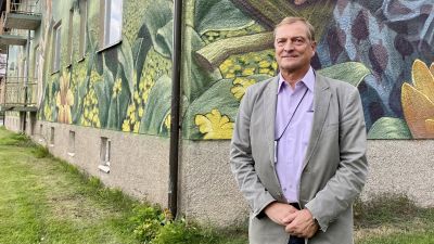 En äldre man står framför en muralmålning på en husvägg. Han ler och tittar in i kameran.