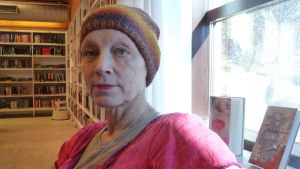 Minna Metzler identifierar sig inte med sin cancer. Minna Metzler i bibliotek. Bokhyllor och böcker i bakgrunden.