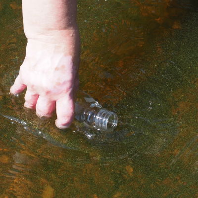 Läpinäkyvään muovipulloon otetaan vesinäytettä rantavedestä jossa on sinilevää.