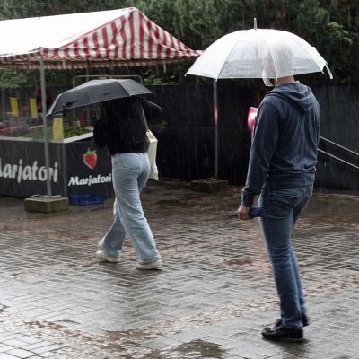 Två personer med paraply över huvudet går på en regnfylld gata i Helsingfors. De går förbi ett bärstånd.