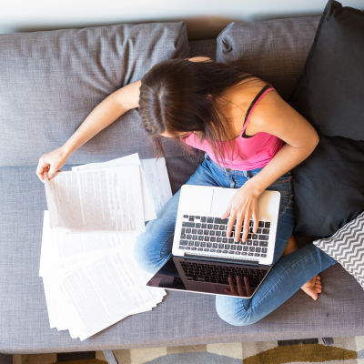 En kvinna sitter i en soffa med sin dator och många papper. Bilden är tagen uppifrån.