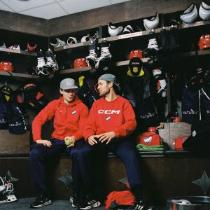Kaksi punapaitaista jääkiekon pelaajaa keskustelevat keskenään pukukopissa. Toisella on kädessään tennispallo. Taustalla on pelikamoja.