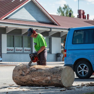 Esko Heikura och de första snitten i träytan. Mies kuulosuojaimet päässä ja moottorisaha kädessä maassa makaavan pöllin äärellä, aloittamassa sahaamista, taustalla parkkipaikka ja kauppa
