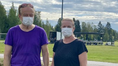 En man och en kvinna med munskydd står framför en gräsplan och tittar in i kameran. Bakom dem syns en scen som håller på att byggas upp.