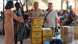 Ingåborna Yrjö Kumpula (till vänster) och Reidar Wilkman från föreningen Schoolkids Light som hjälper skolbarn i Tanzania. Några Ingåbor brukar varje vinter resa till Tanzania för att säkra sig om att all hjälp kommer fram till rätta adresser. Två (vita) män står vid en stor hög med lådor och andra förpackningar tillsammans med några afrikanska kvinnor. I bakgrunden en massa barn som sitter och följer med. Platsen är i en skola i Tanzania i Afrika.