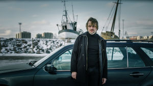 Usko Bergdahl (Jani Volanen) seisoo autonsa edessä lumisessa maisemassa. Taustalla kaksi suurta venettä ja kaupungin siluettia.