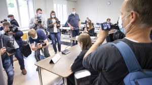 På grund av coronaviruset ska alla som befinner sig i rättssalen bära ansiktsskydd. Den åtalade i skolattacksfallet i Kuopio sitter med ryggen mot kameran i en rättssal. Flera fotografer står framför honom och fotograferar. En fotograf syns på bild som står bakom mannen. Samtliga personer har munskydd på sig.