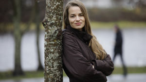 Matilda Sorkkila säger att mammor upplever att de har stora krav när det kommer till att vara förälder. Kvinna lutar sig mot ett träd med armarna i kors och ler.
