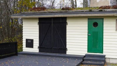 En stransbastu med ljusbeige fasad, svarta skjutdörrar och en grön dörr. Bastun har grästak. På fasaden finns en solfångare monterad.