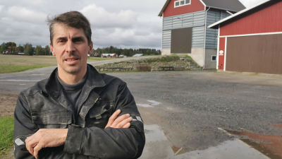 Mansperson i arbetskläder står utomhus, i bakgrunden rödmålade lantbruksbyggnader.