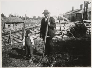 Mies ja poika maatilan pihalla 1900-luvun alussa, miehellä sirppi kädessä ja viikate olalla