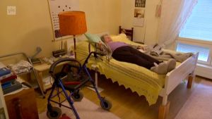 Äldre kvinnan Hilkka Jämsä ligger i en säng med rollator bredvid sig.