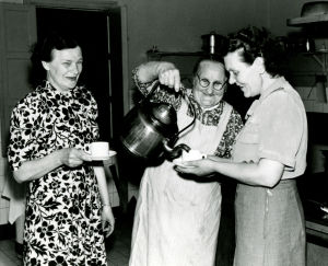 Kökshjälpen Amalia Tiltti häller upp en skvätt kaffe åt kassörskan Kerttu Liukkonen (t.v.) och servitrisen Aune Nieminen (t.h.). Bilden är från Lahtis stationsrestaurang, 1952. Tre kvinnor på rad, den mittersta häller upp kaffe ur en enorm kopparpanna.