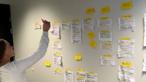 Henkilö pitää post it-lappua kädessä taulun edessä, taululla muita lappuja.