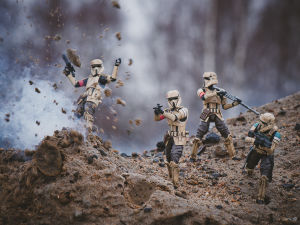En bild med en praktisk effekt. Fyra leksaksfigurer föreställande soldater från Star wars. De står på sand om en av dem verkar ha stigit på en mina. Det sprättar upp sand framför honom.