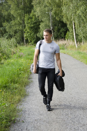 En man iklädd svarta jeans och en grå t-skjorta går på en grusväg kantad med grönt gräs och blommor. Han ler och ser glad ut.
