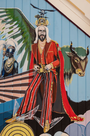 Jeesus kuvattuna punaisessa puvussa, raidalliset housut, kädessä nuottiavain, lahkeessa kolme kahdeksaa, takin liepeessä usko, toivo ja rakkaus