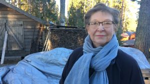 Tuula Alkias sommarstuga ligger vid havet ungefär tre kilometer från flygplatsen. Medelålders kvinna tittar in i kameran. Bakom henne synns träd och en vedbod.