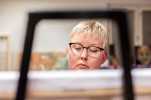 Lähikuvassa Elina Lehtoranta maalaustelineen takana. Katsoo hieman alaviistoon.